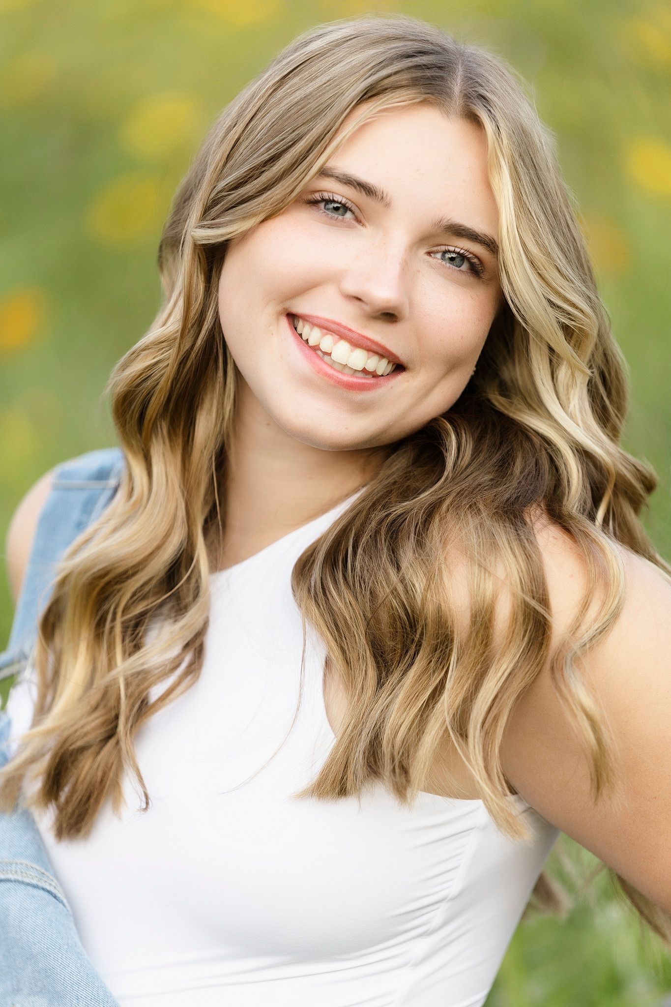 A bright outdoor portrait showing a natural smile and shoulder-length wavy hair against a green bokeh background.