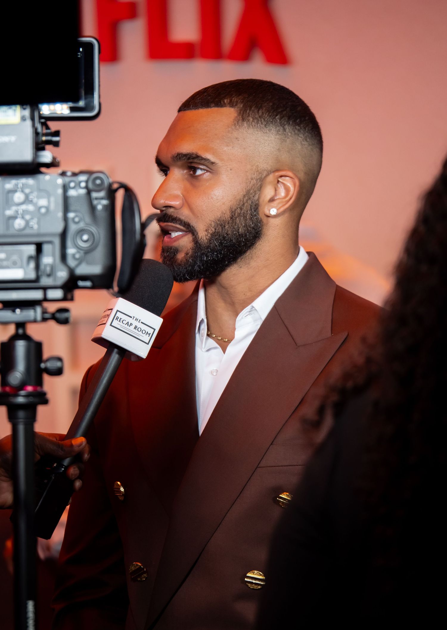 Person in brown suit being interviewed against a red Netflix backdrop with cameras visible.