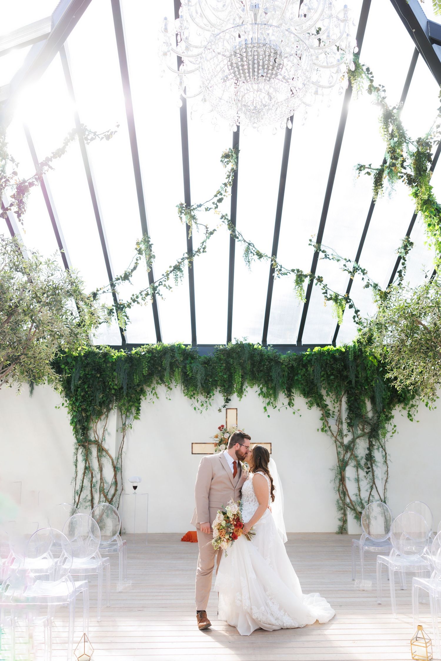 A couple shares an intimate moment under a crystal chandelier and greenery-adorned glass ceiling in an elegant wedding venue in Branson.