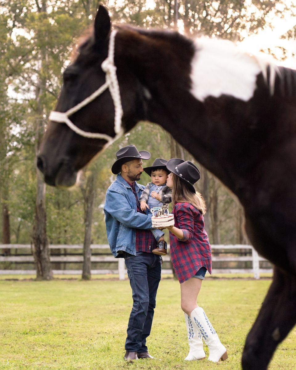 Couple hugging and celebrating outdoors with a horse in field near trees and white fence during a family photoshoot in Gables