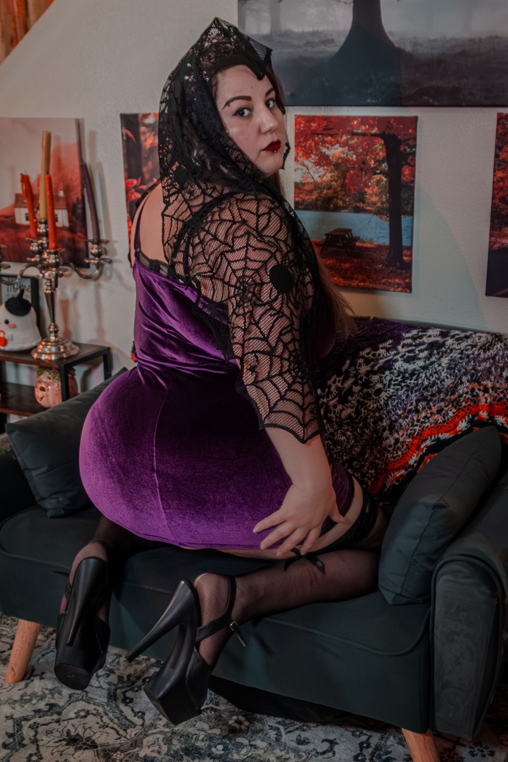 A figure in a purple velvet dress and black lace spider web headpiece poses on a grey couch in a moody Halloween setting.