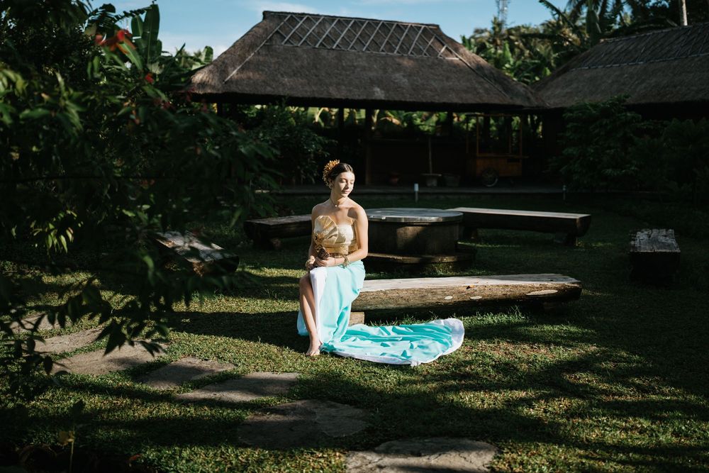 Traditional tropical photoshoot in garden setting with teal sarong against rustic architecture.