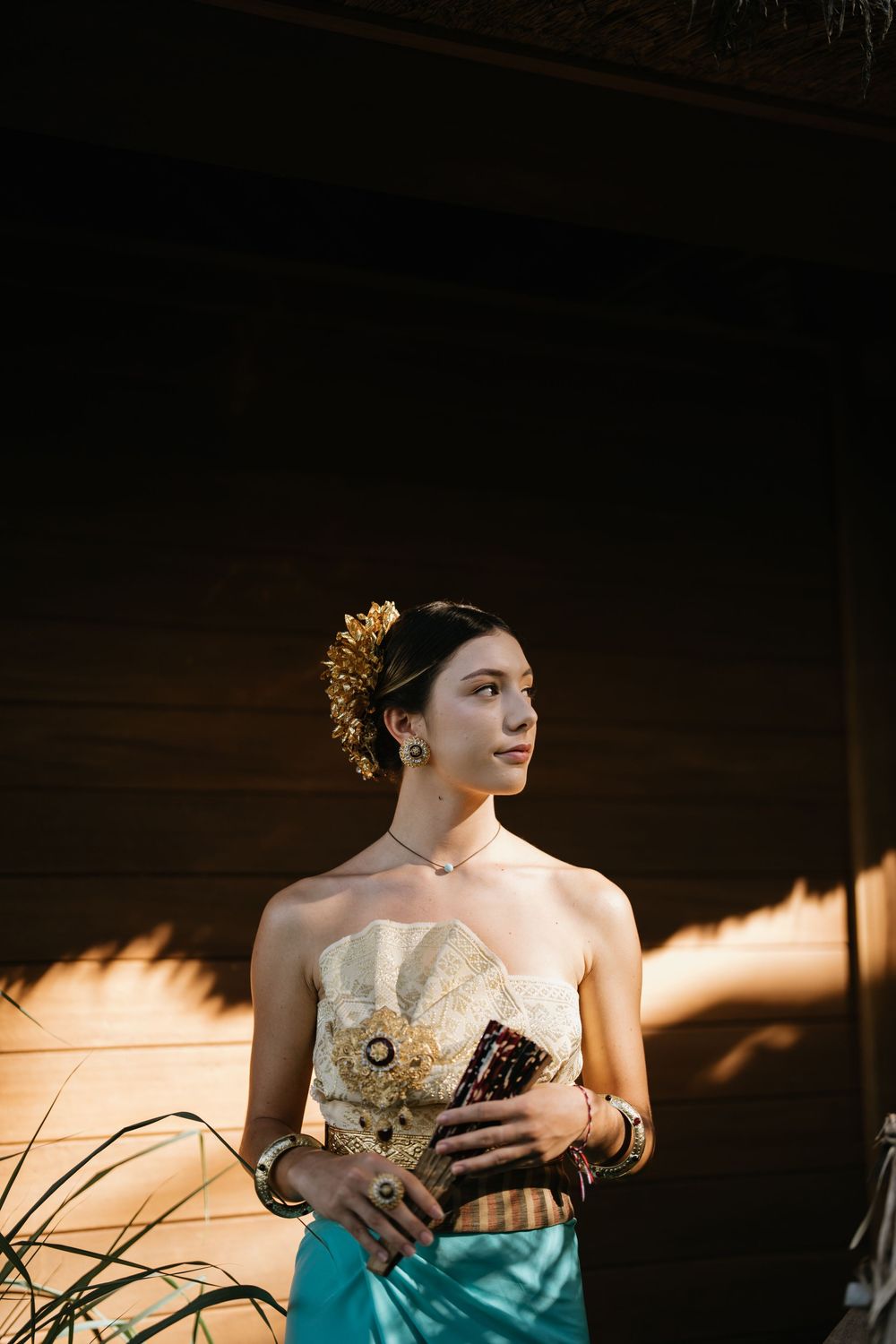 Woman in traditional Balinese-inspired outfit with gold hair accessories and cream corset standing in warm sunlight.