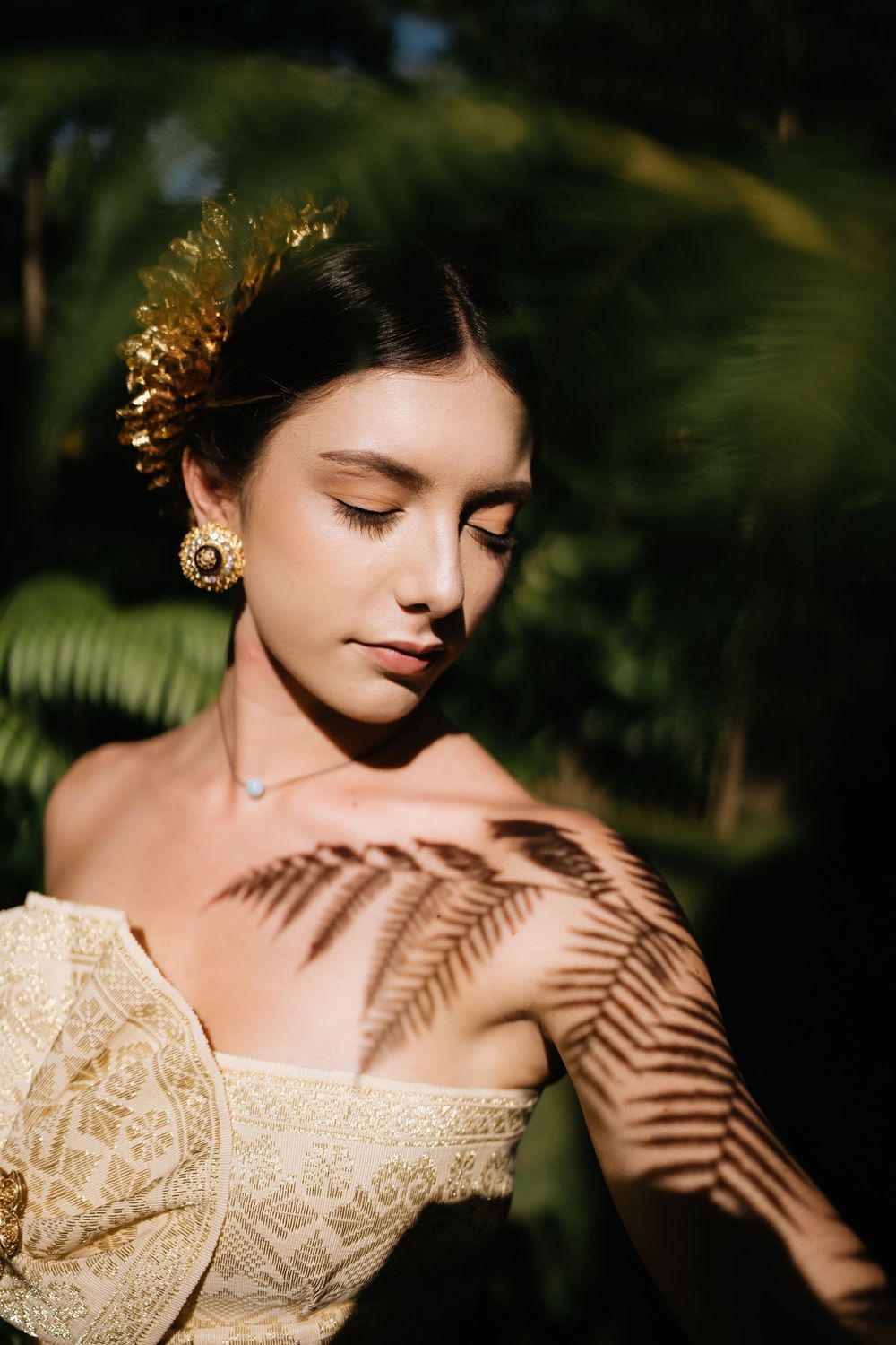 Artistic portrait with fern shadow patterns and gold hair accessories in natural lighting.