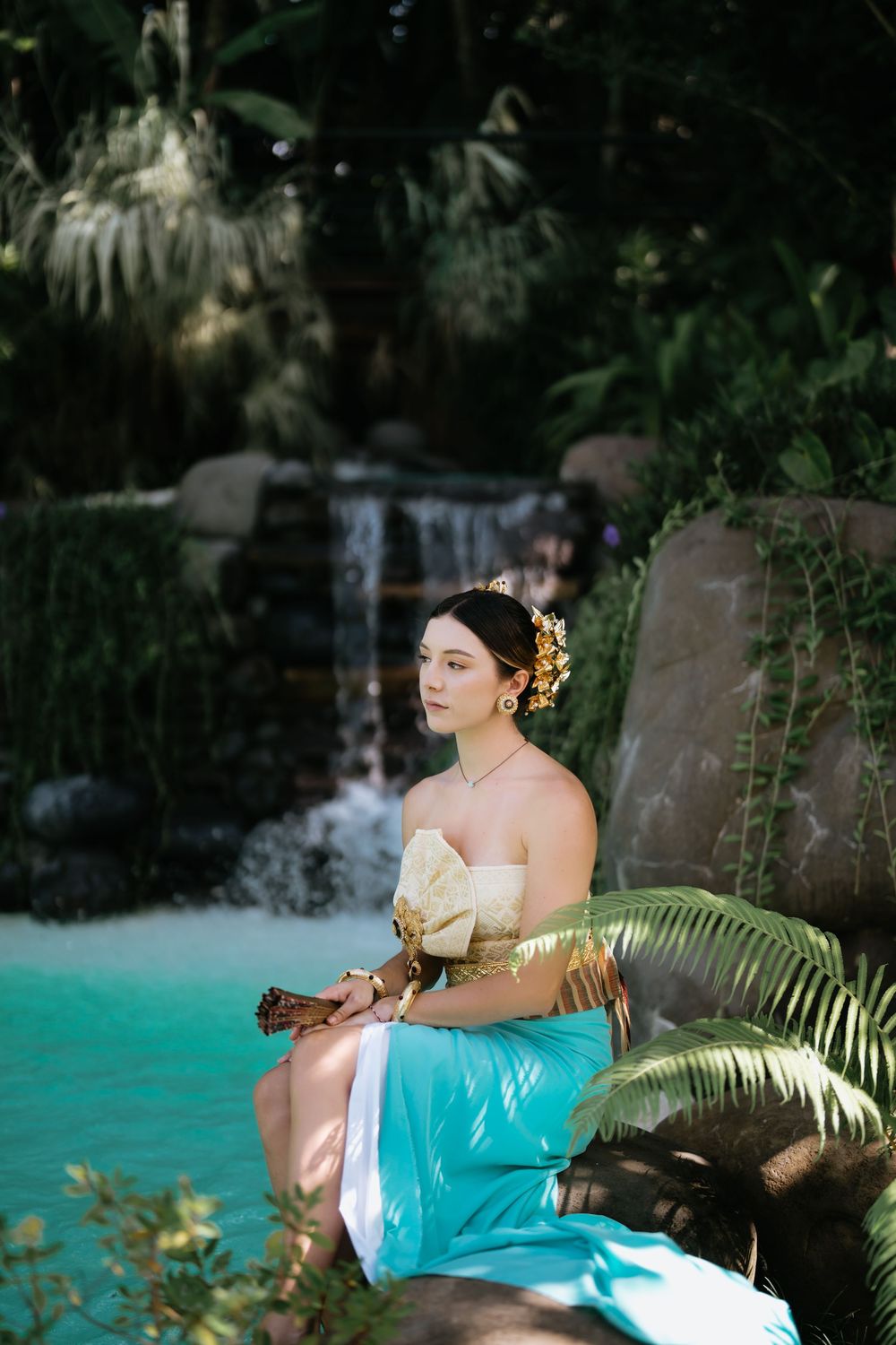 A traditional Thai-inspired photoshoot by a turquoise pool with a waterfall and tropical foliage in the background.