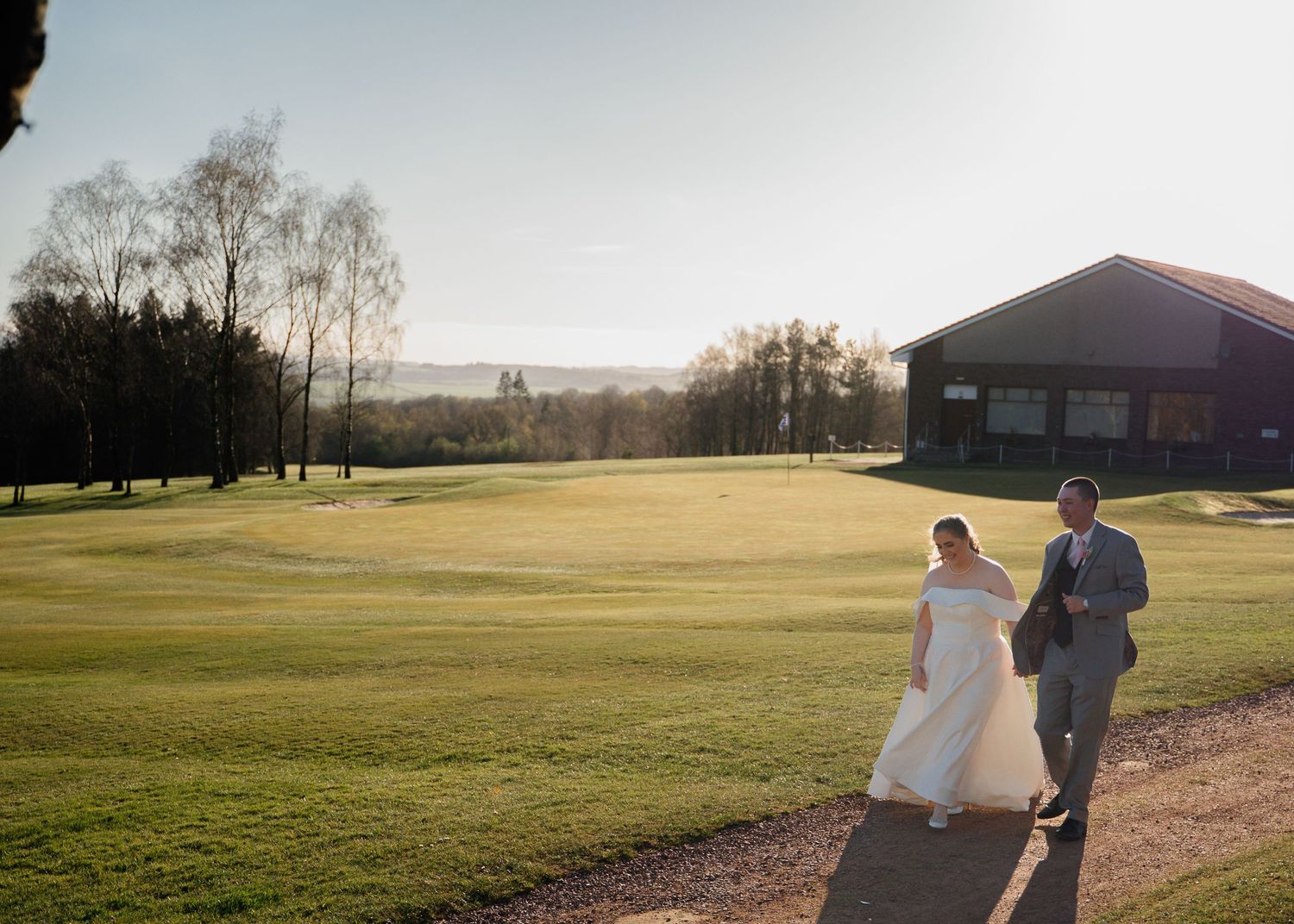 A couple walks along a scenic golf course path at sunset with a brick building in the background.
