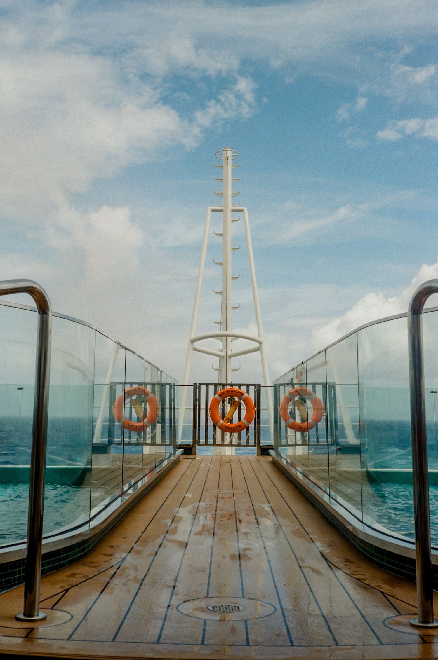 Wooden deck walkway on a cruise ship with glass railings, life preservers, and a white mast against blue sky and ocean.