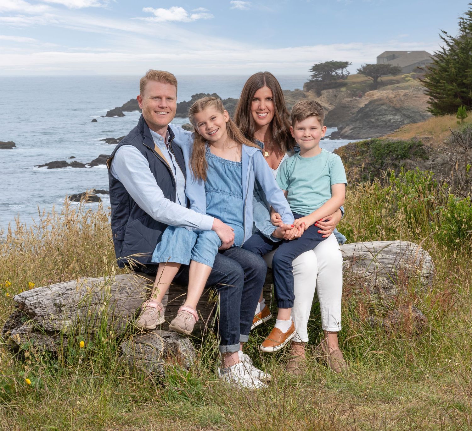 A group of people relaxes together on a grassy bluff overlooking the Pacific Ocean on a sunny afternoon.