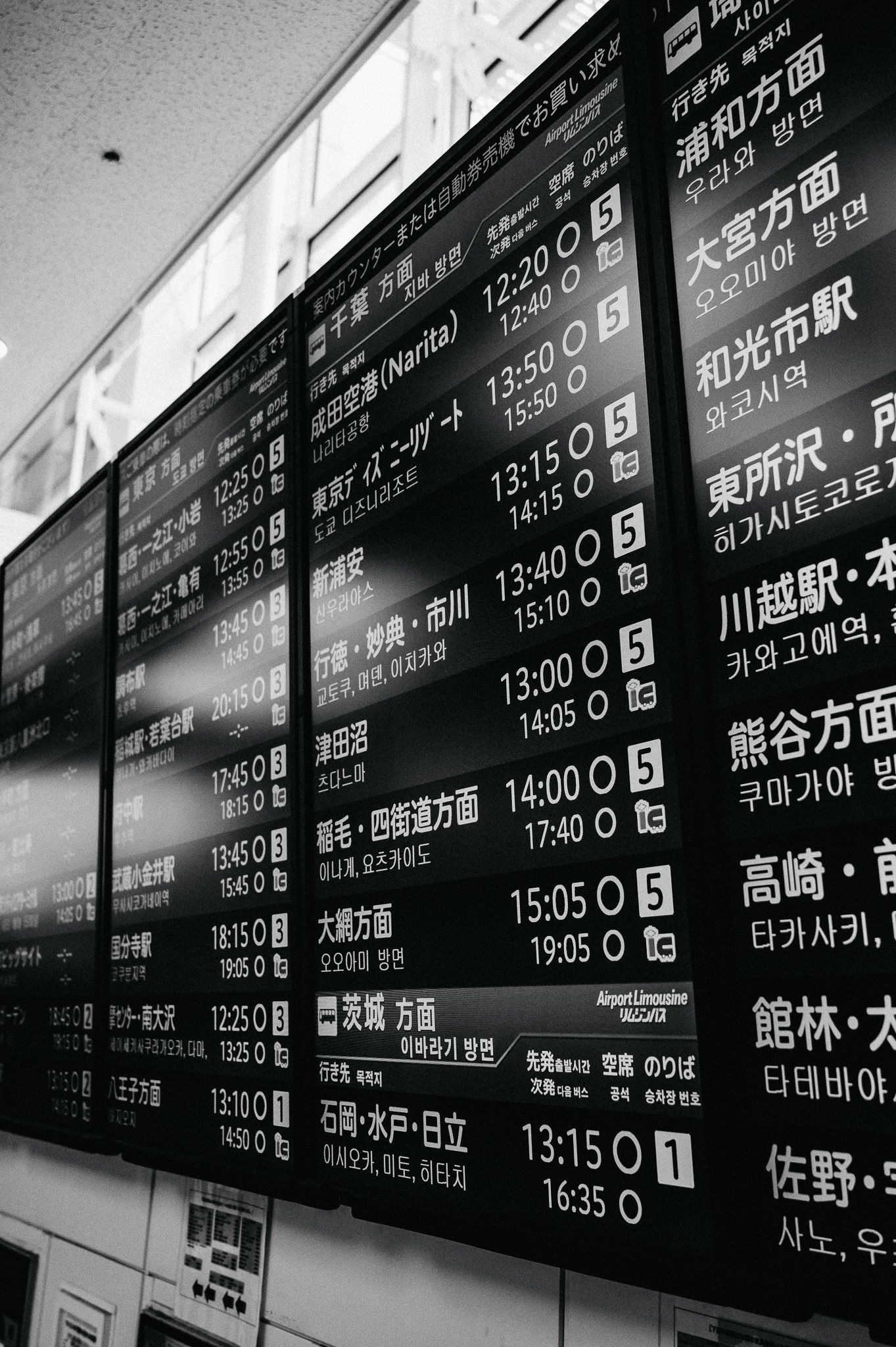 Black and white photo of Japanese train station departure board with arrival times and destinations in multiple languages.