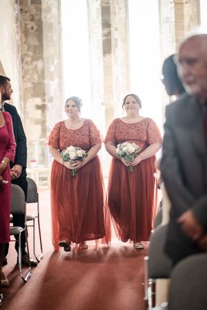 Two bridesmaids in matching rose-colored gowns hold bouquets during a traditional church wedding ceremony.