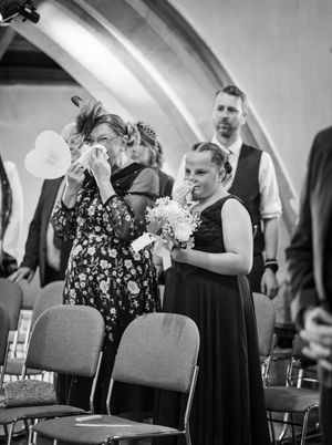 Black and white photo of two wedding guests at a church ceremony sharing an emotional moment among folding chairs.