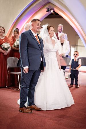 At a wedding ceremony, the bride in a white gown stands and dances with a guest wearing a navy suit on red carpeting.