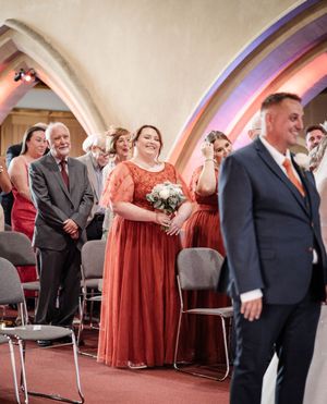Wedding guests in burgundy dresses and formal suits stand in an arched church setting with warm lighting.