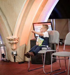A young child sits on a chair while pointing at something in a room with curved architectural details and soft lighting.