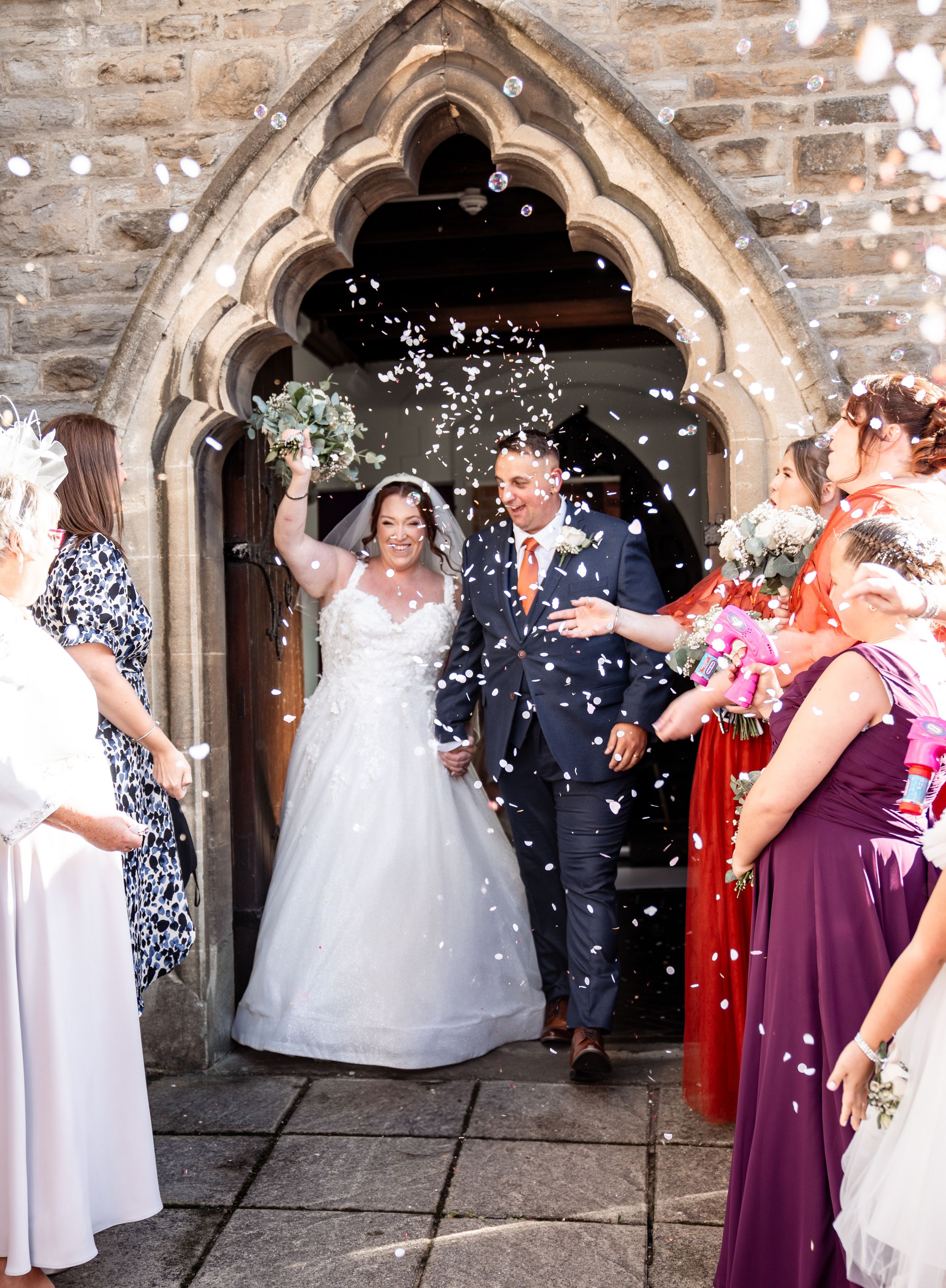 Wedding party celebrates with confetti outside a gothic stone church archway during a joyous celebration.