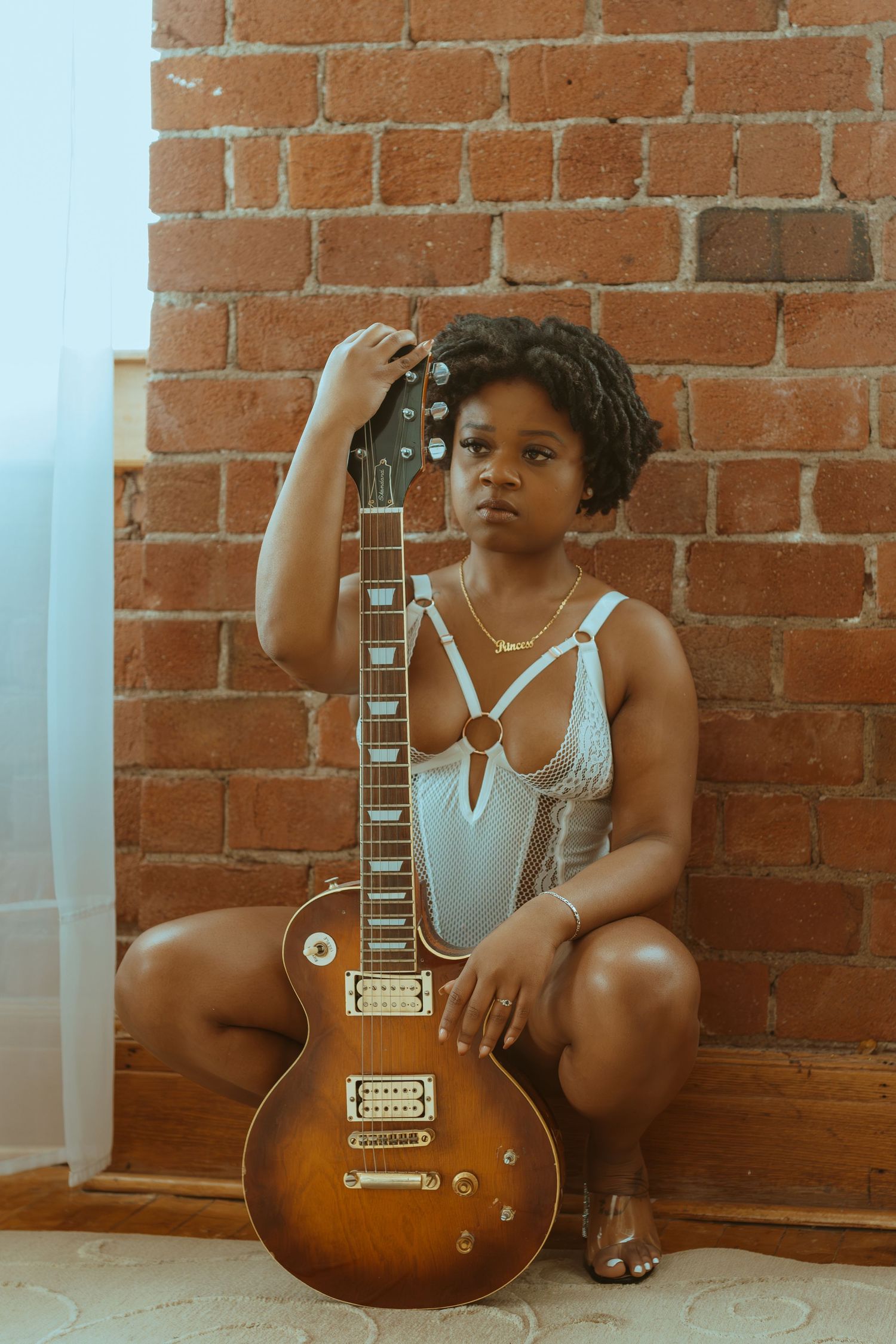 Figure shown in casual white tank top poses with a vintage sunburst electric guitar against exposed brick wall.