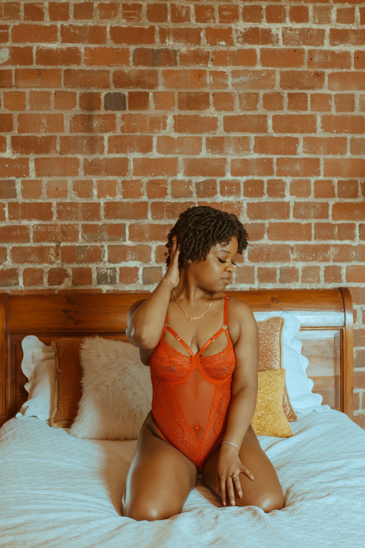 Figure reclining on bed with pillows wearing orange lingerie against rustic brick backdrop.
