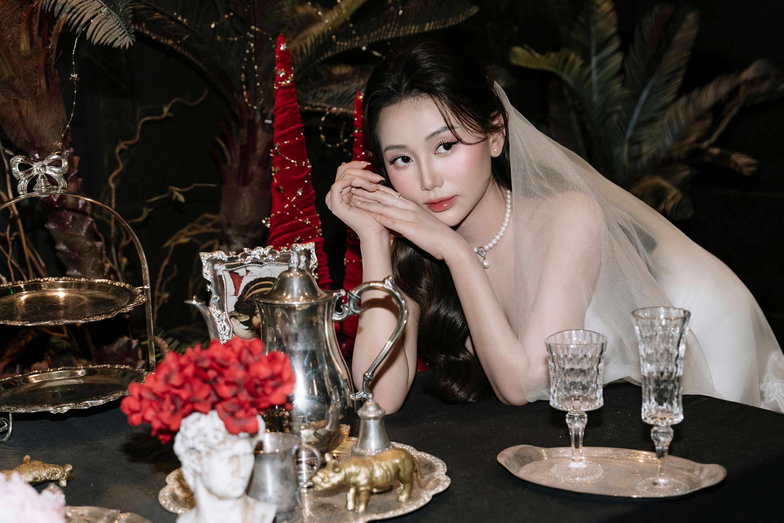 Bride in white wedding dress poses elegantly at vintage table with crystal glassware and roses.