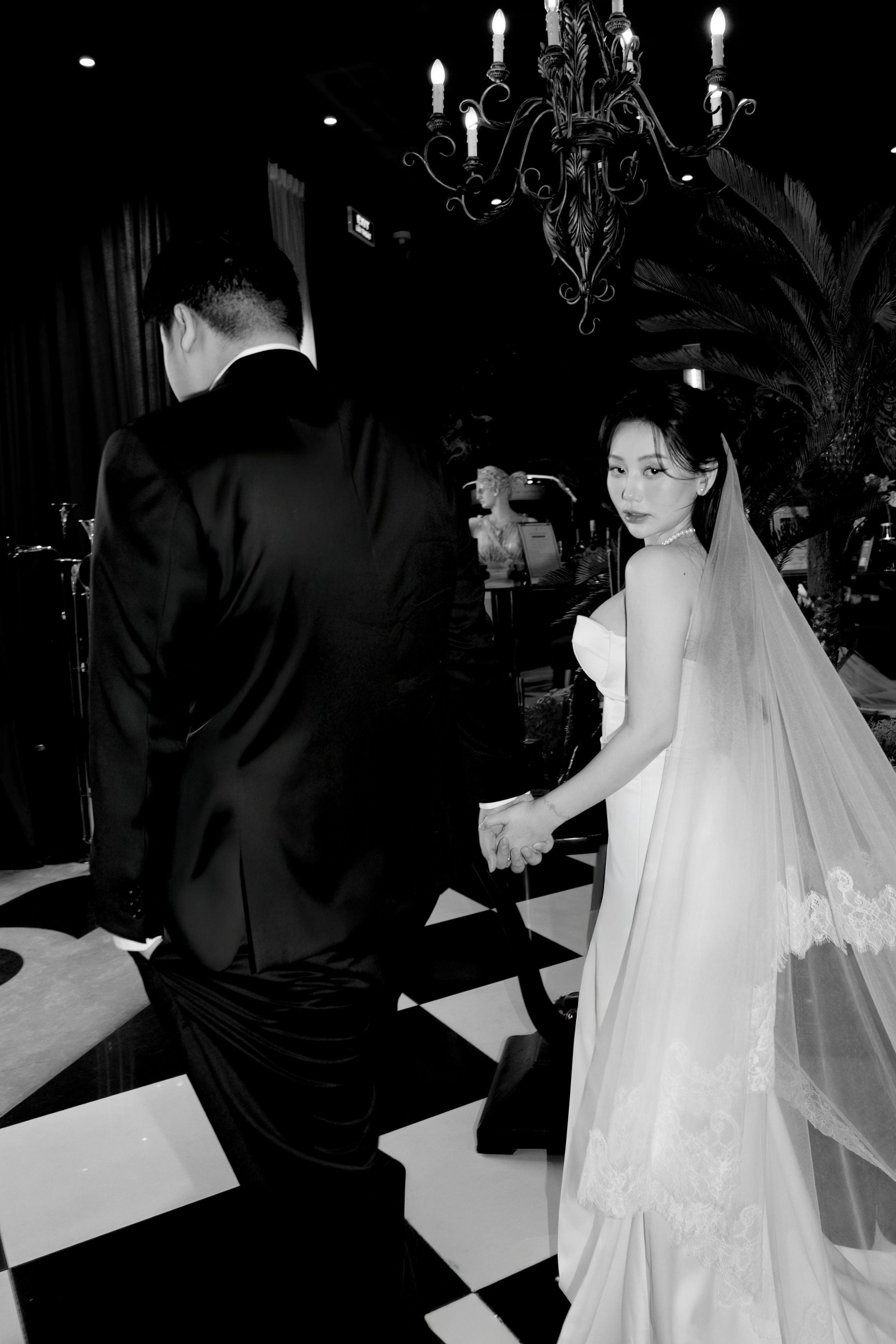 Black and white photo of bride and groom holding hands walking on checkered floor at wedding.