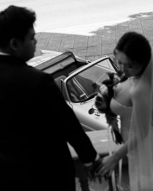 Black and white candid photo of bride holding bouquet beside groom near wedding car outside.
