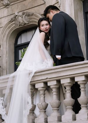 Bride in lace wedding gown with long veil leans on balcony railing with groom in black suit.