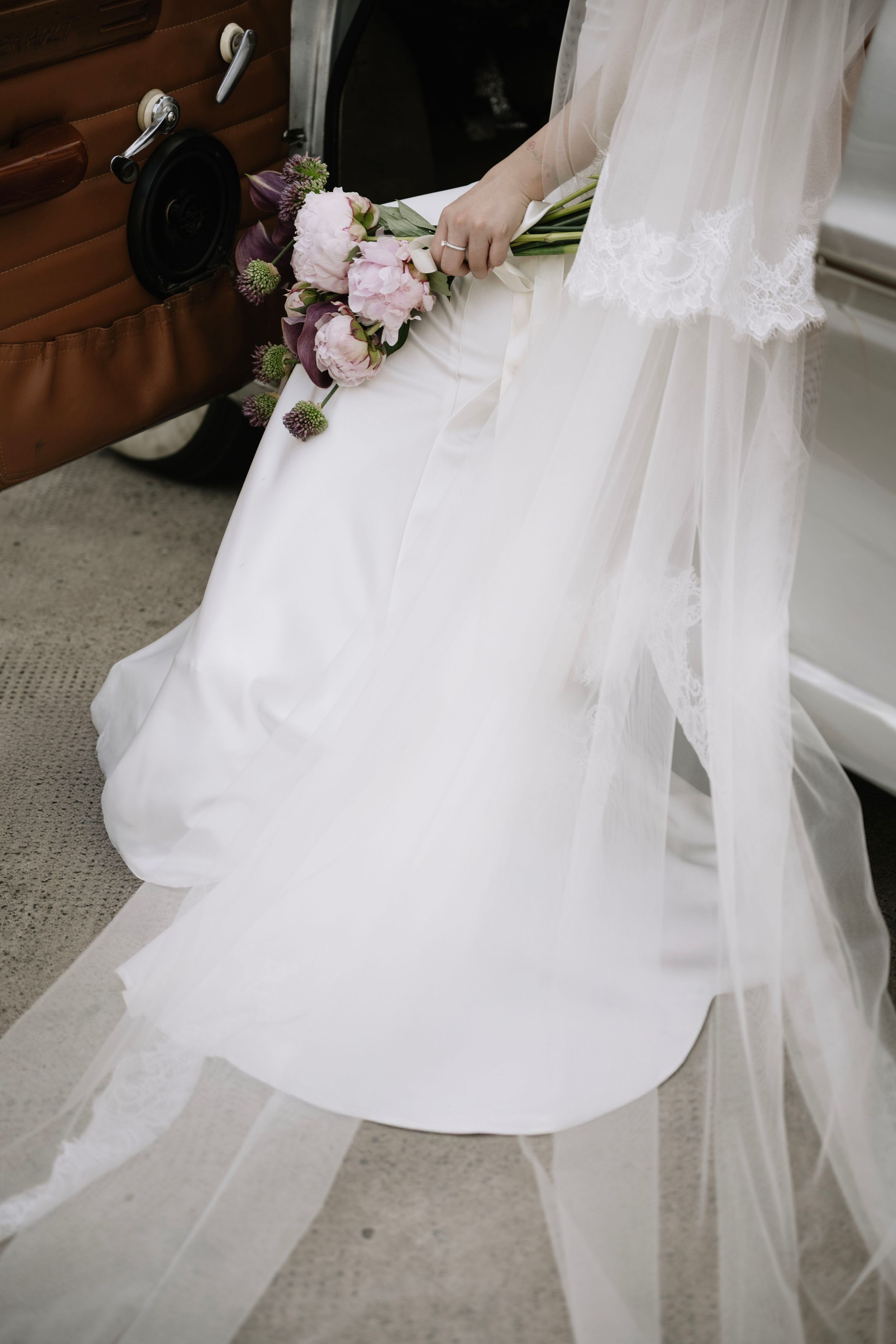 Bride in white lace wedding gown holding pink and purple floral bouquet beside vintage brown car.