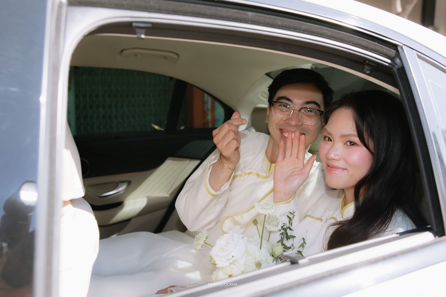 Newlywed Vietnamese couple in white ao dai wave and smile from inside wedding car.