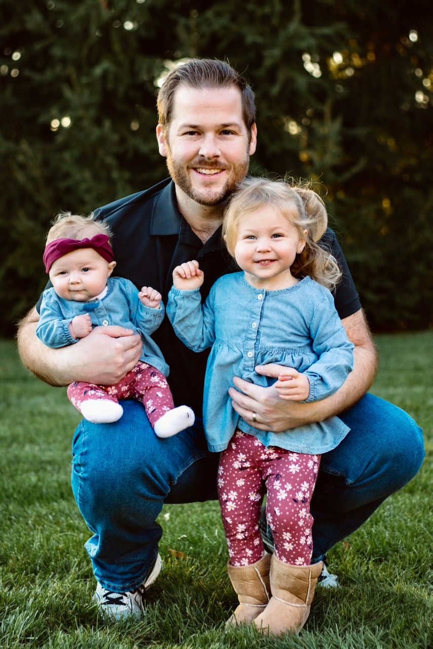 Father keeling with his two daughters smiling during a lifestyle family photography golden hour session in South Lyon, Michigan.