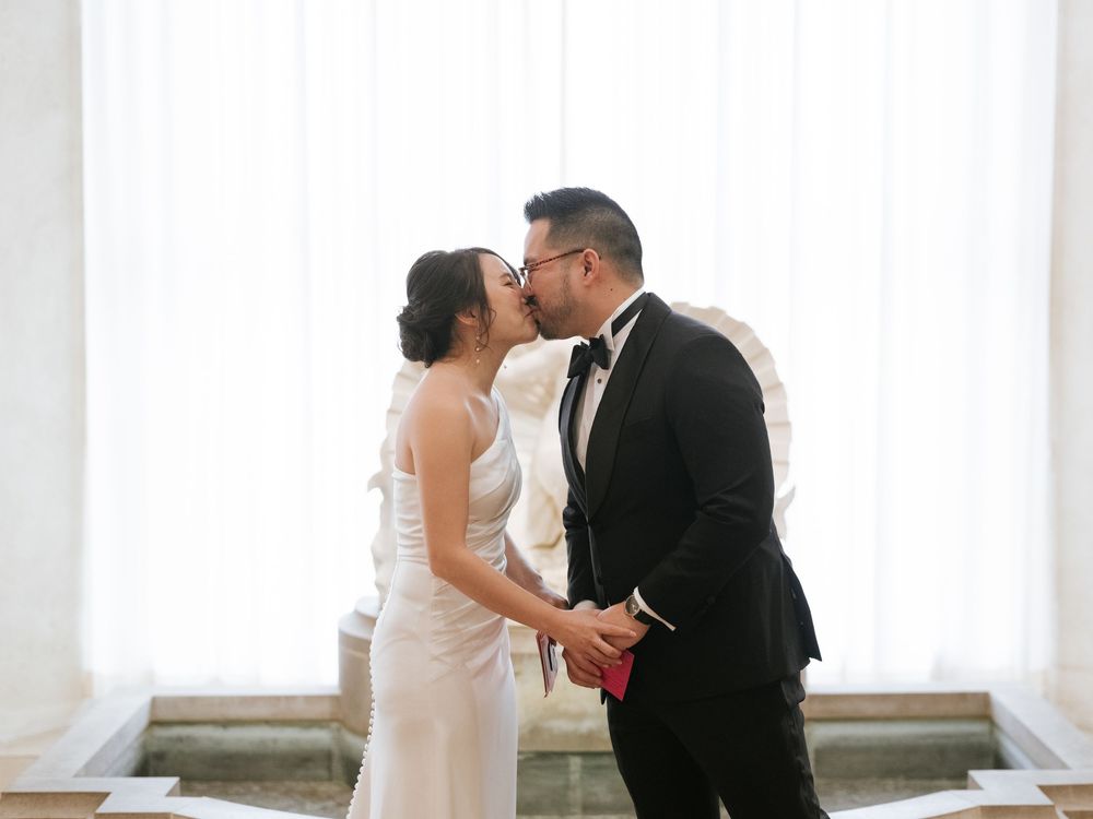 A couple in wedding attire sharing an intimate moment in a bright, airy room with white curtains.