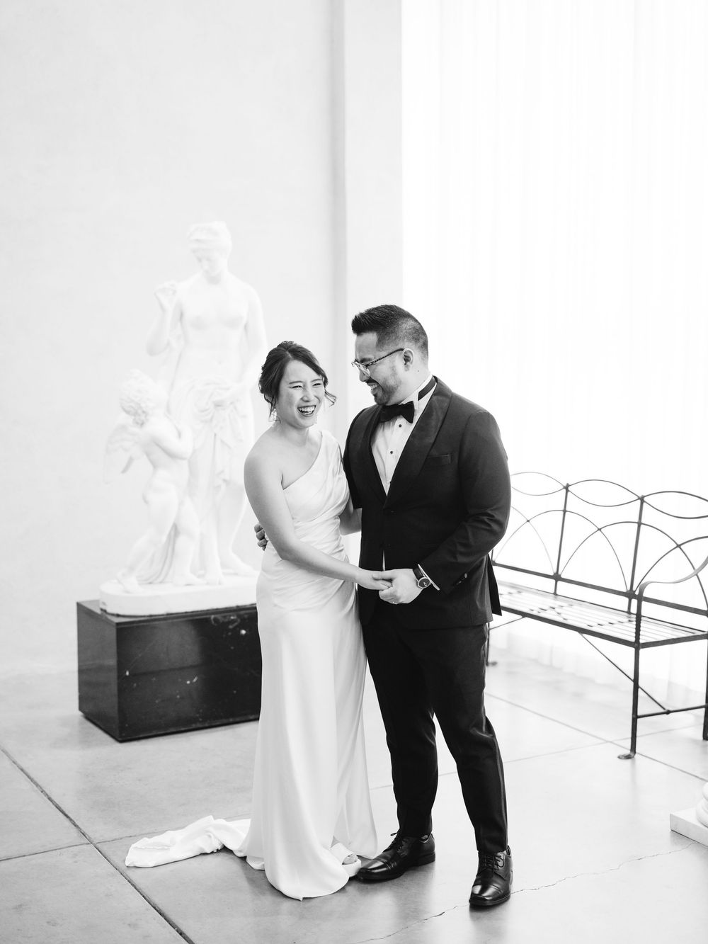 A couple shares a tender moment in a minimalist white room with a black trunk visible in the background.