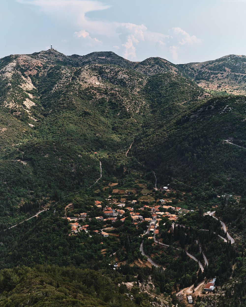 Panoramic mountain landscape showing winding roads and scattered buildings among green Mediterranean hills.