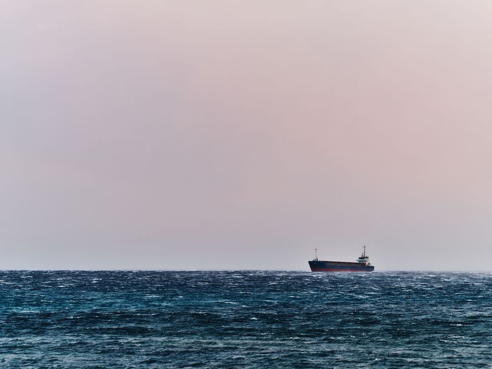 Cargo ship silhouetted on horizon against pastel sky and deep blue ocean waters.