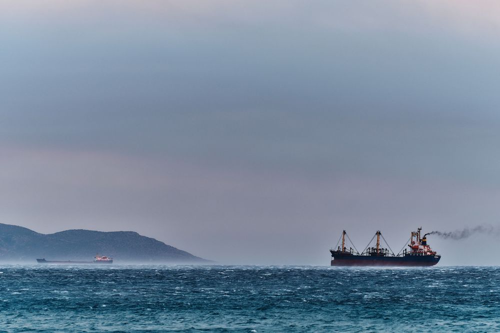 Dramatic seascape with dark stormy clouds and a cargo ship silhouetted against a moody ocean horizon.