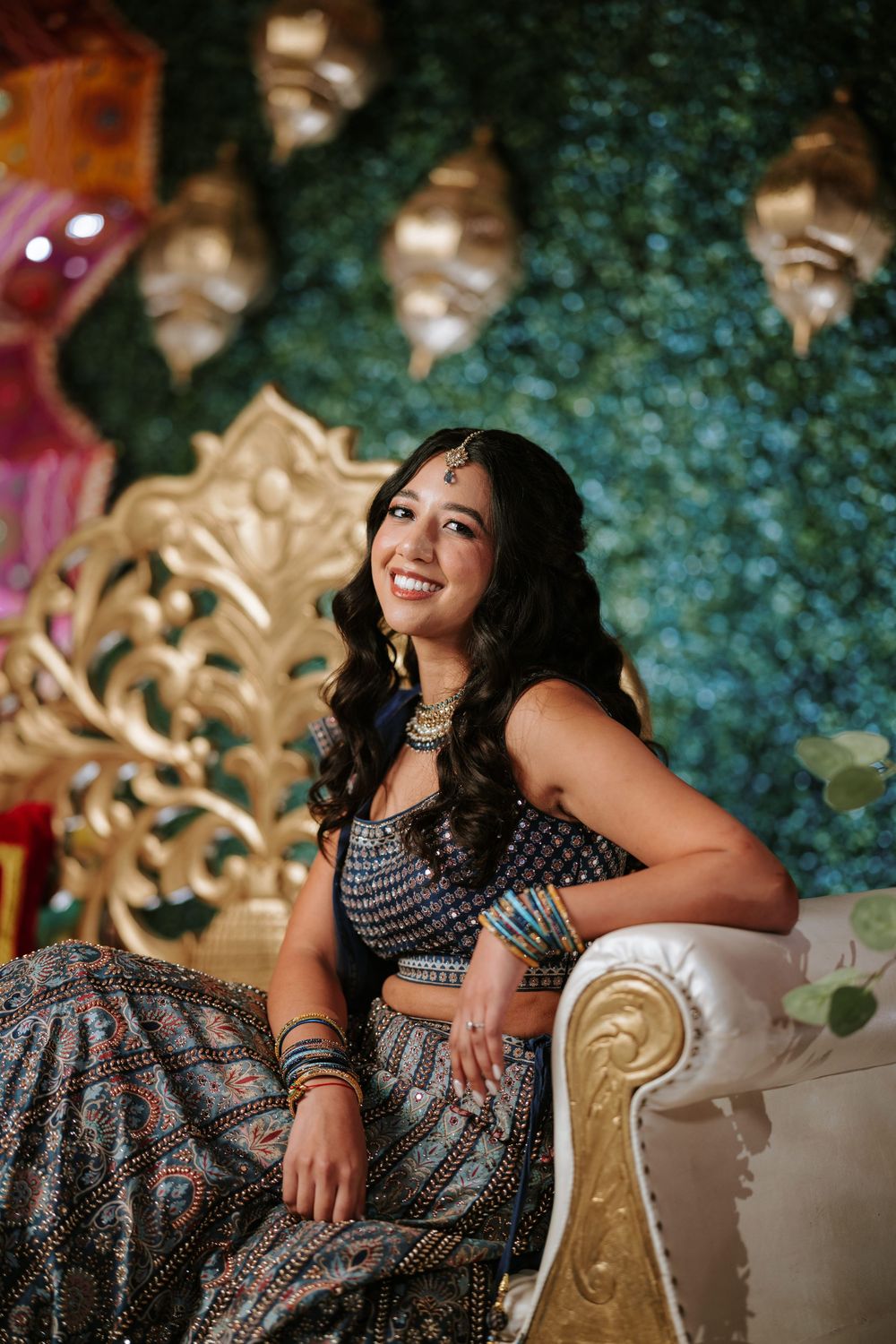 A woman in a black and white patterned dress poses elegantly on an ornate chair against a floral backdrop.