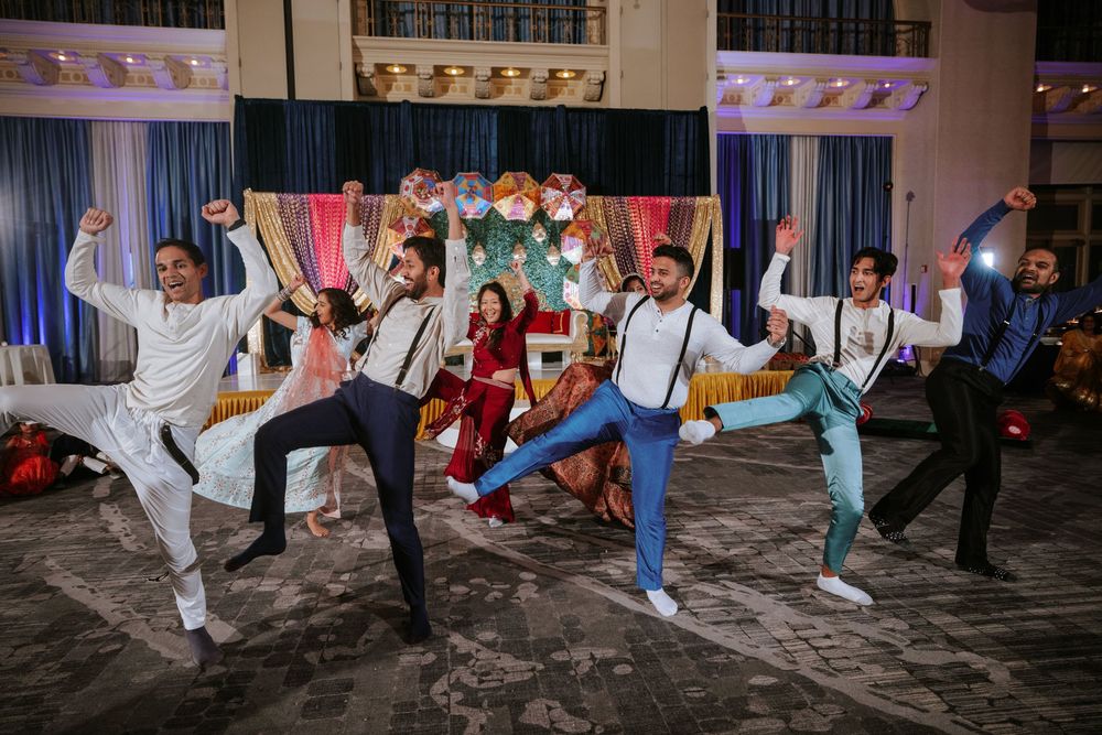 Group of wedding guests perform synchronized dance moves wearing suspenders and colorful outfits.
