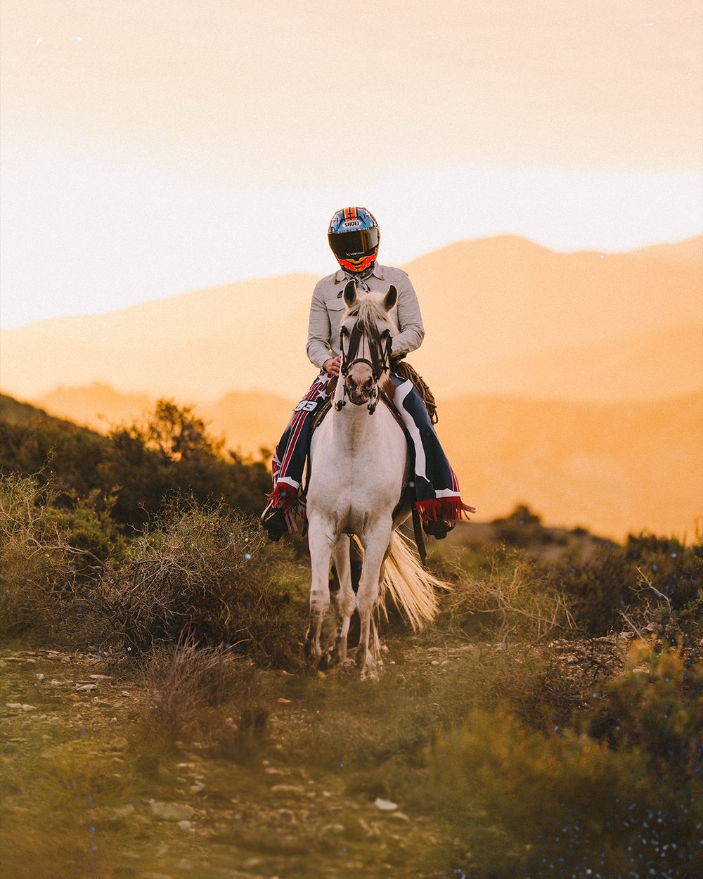 A rider guides a white horse through golden desert terrain at sunset against mountain silhouettes.