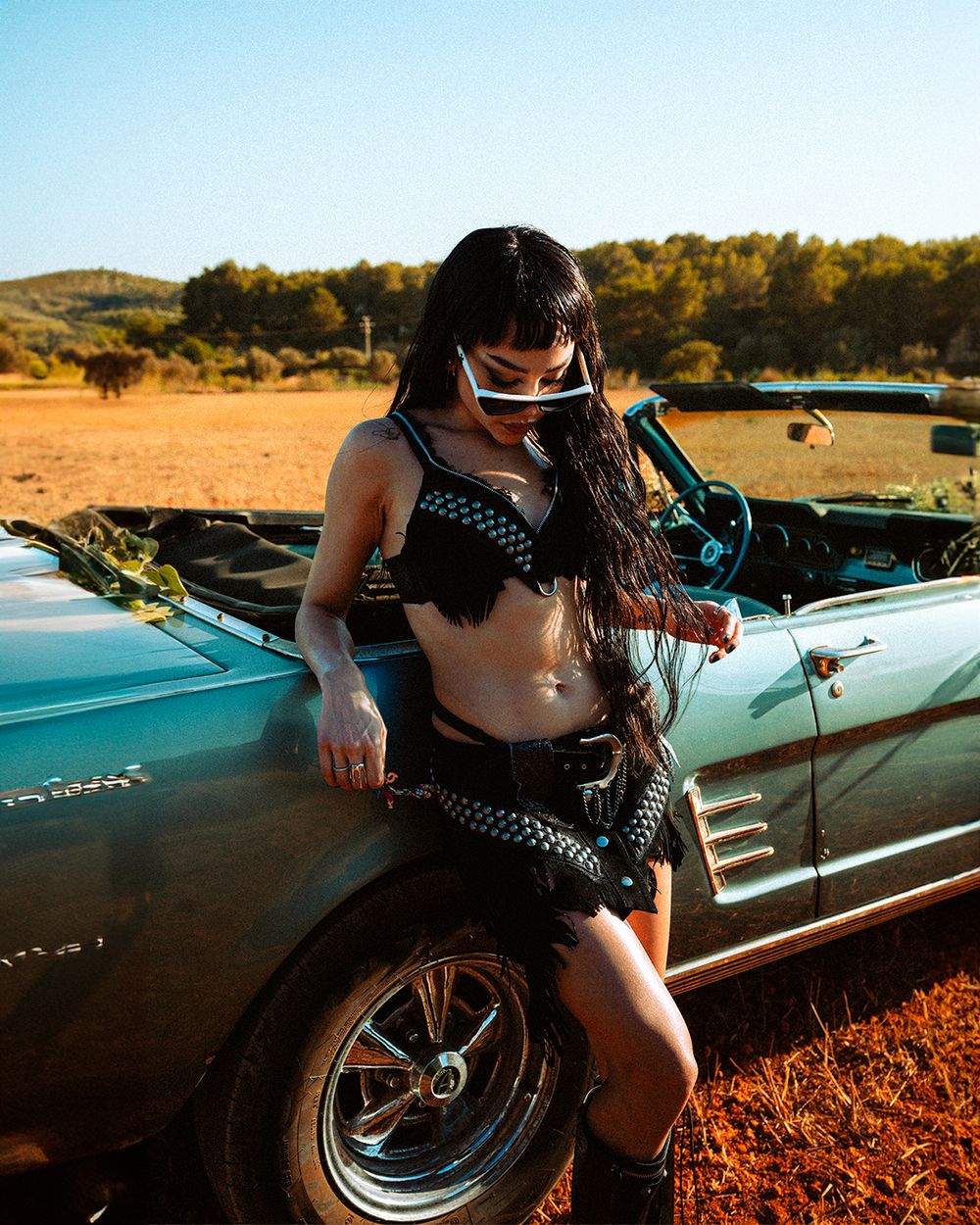 A person in punk rock style clothing poses next to a classic car on dusty ground during golden hour.