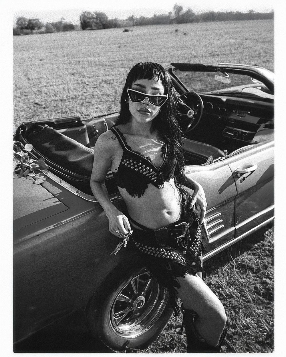 Vintage black convertible car parked in grassy field with retro-styled model posing in polka dot outfit.