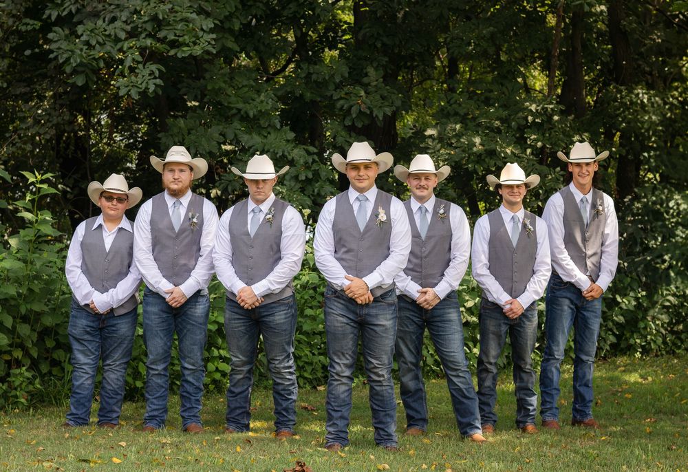 A row of seven groomsmen in matching western attire with cowboy hats, gray vests, and blue jeans stand outdoors.