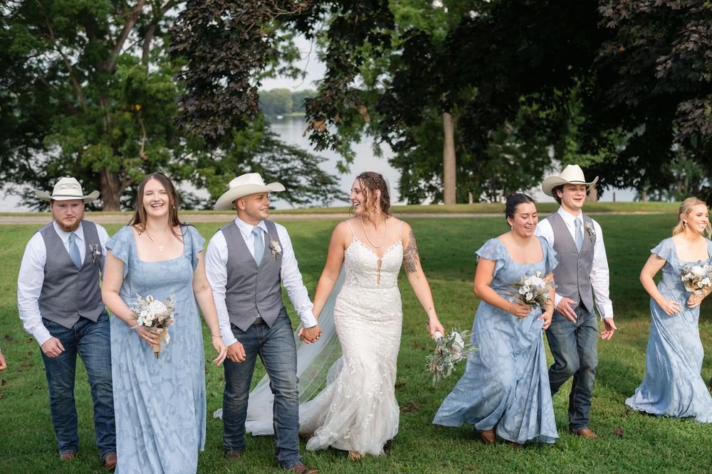 Wedding party in light blue and gray attire poses playfully on green lawn for group photos.