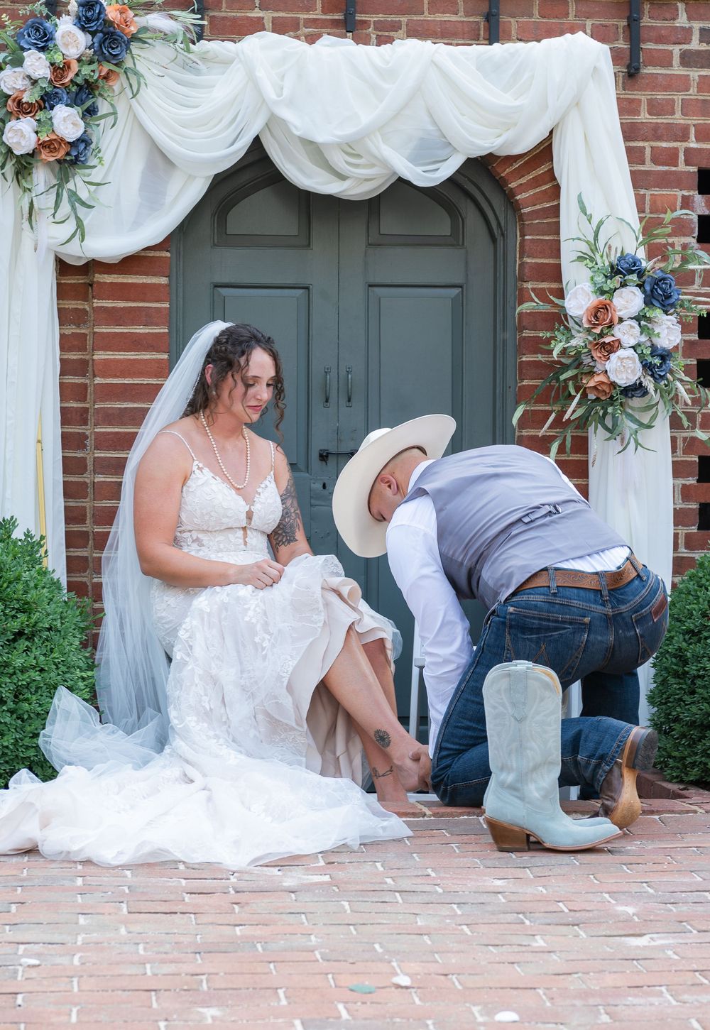 Wedding party members help put on white cowboy boots on a brick patio during an outdoor ceremony.