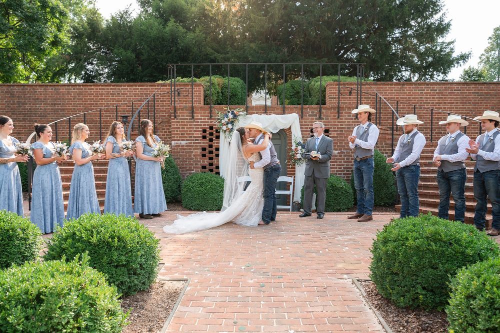 Wedding party stands in line on brick patio with greenery and garden backdrop during outdoor ceremony.