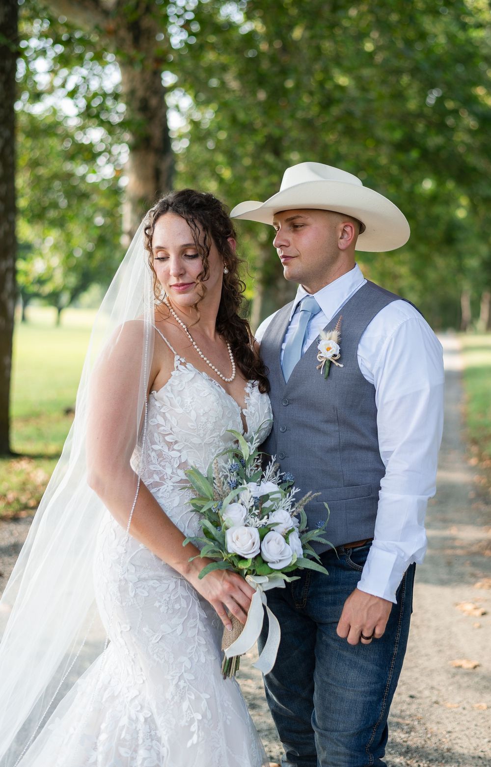 A romantic wedding photoshoot on a tree-lined path shows a couple in western attire with a white dress and cowboy hat.
