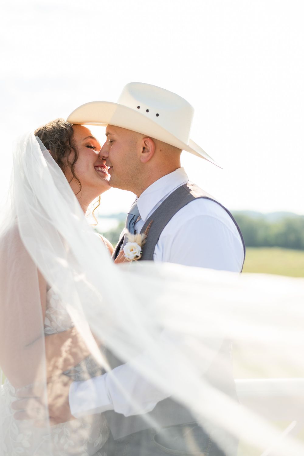A romantic wedding couple shares a tender kiss under a flowing white veil on a sunny day.