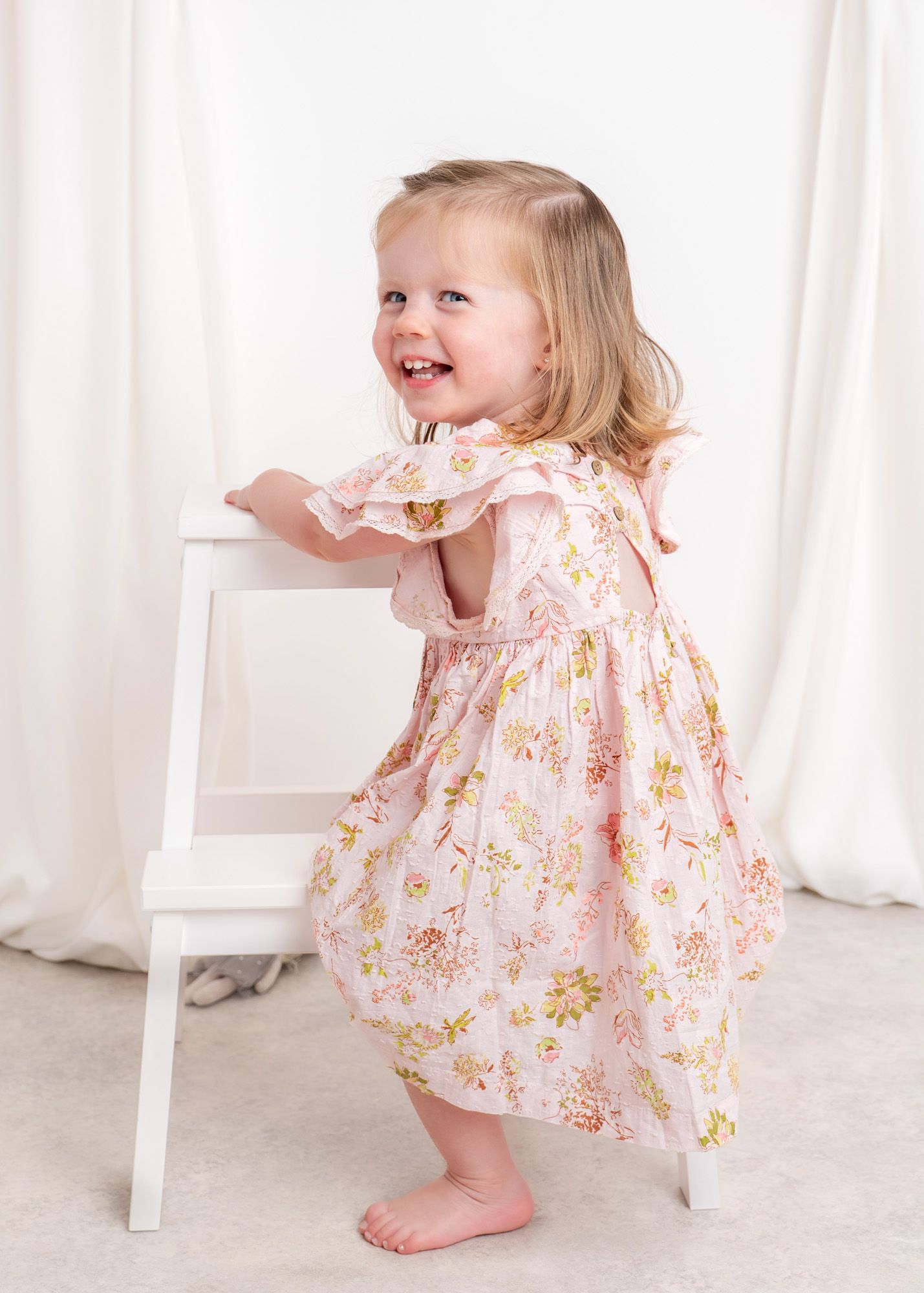 Toddler in floral pink dress smiles while holding onto white chair at birthday session.