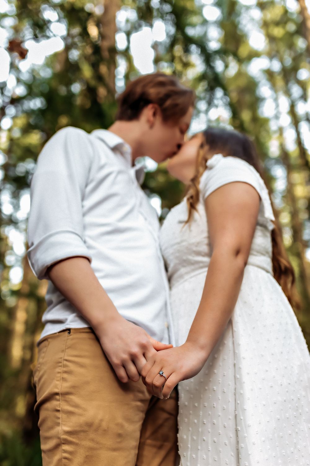 Close-up shot of a couple's tender embrace in white and khaki attire against a forest backdrop.