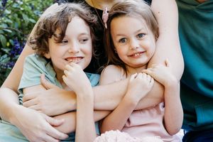 Two young children being embraced in a loving pose outdoors against green foliage.