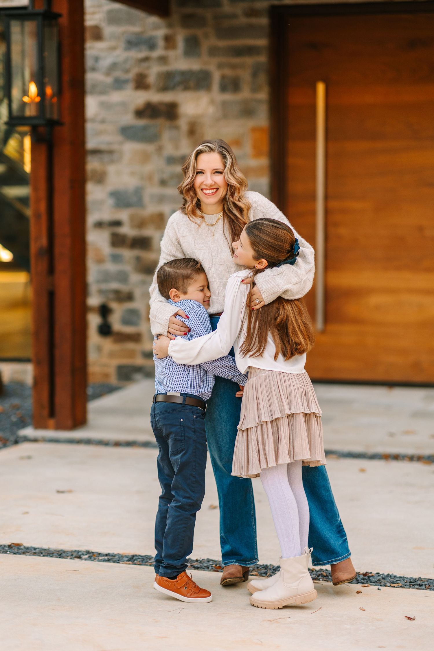 A parent and children share a warm embrace outside a modern stone and wooden entrance.