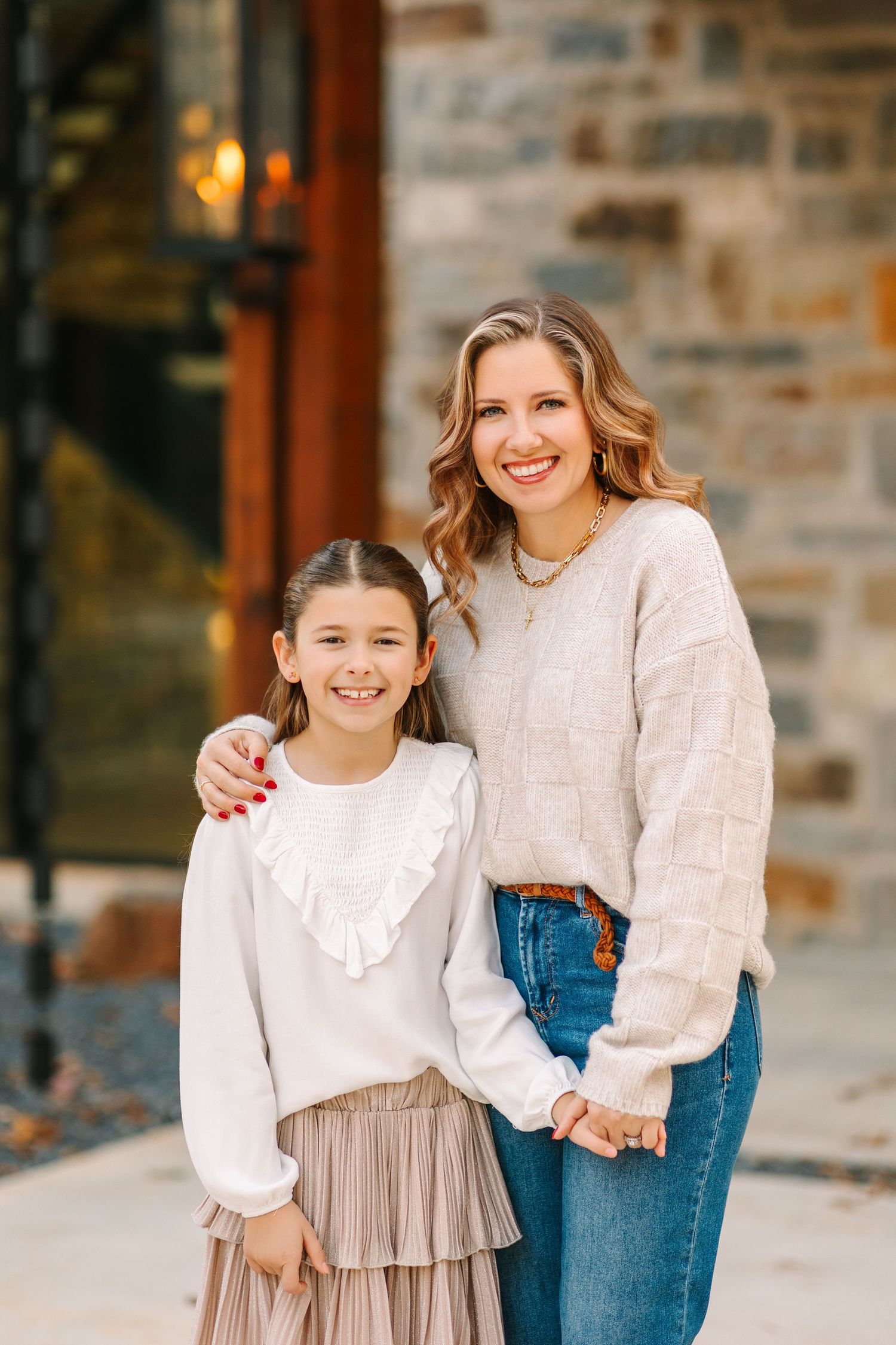 Mother and daughter in matching white sweaters share a joyful embrace while posing outdoors against stone wall.