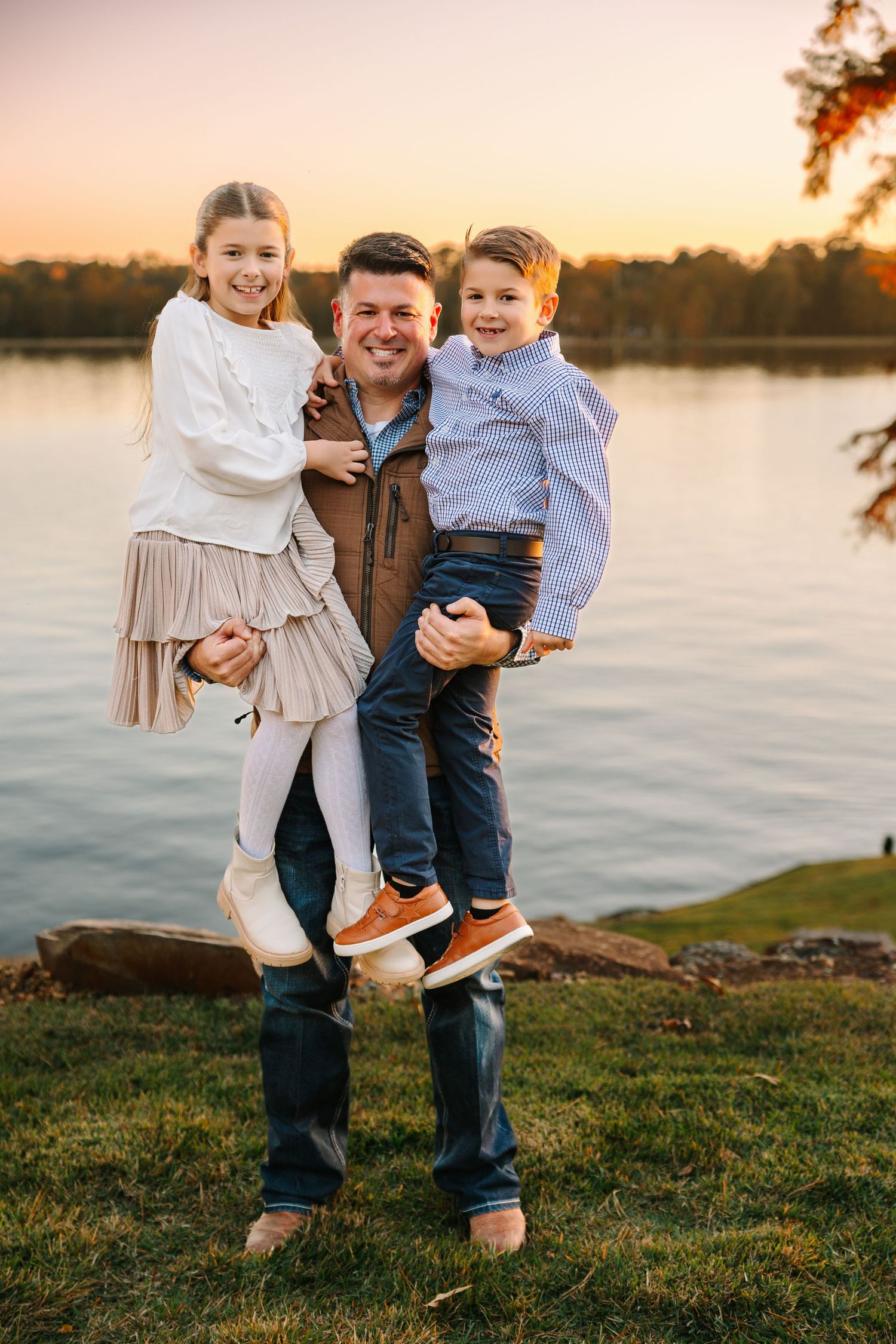 Happy family sharing playful moments together by the lake at sunset with water and grass in background.